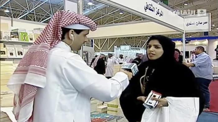 A man dressed in eldishdasha, the long white robe worn by men on the peninsula, hands the microphone to a young woman dressed in an abaya.